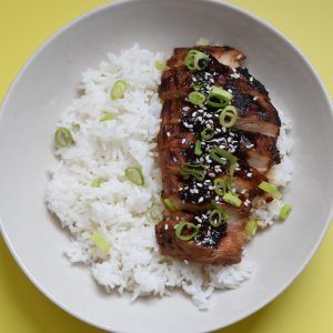 On a yellow backdrop, A plate of rice and honey soy chicken breast sliced up and garnished with sesame seeds and green onions.