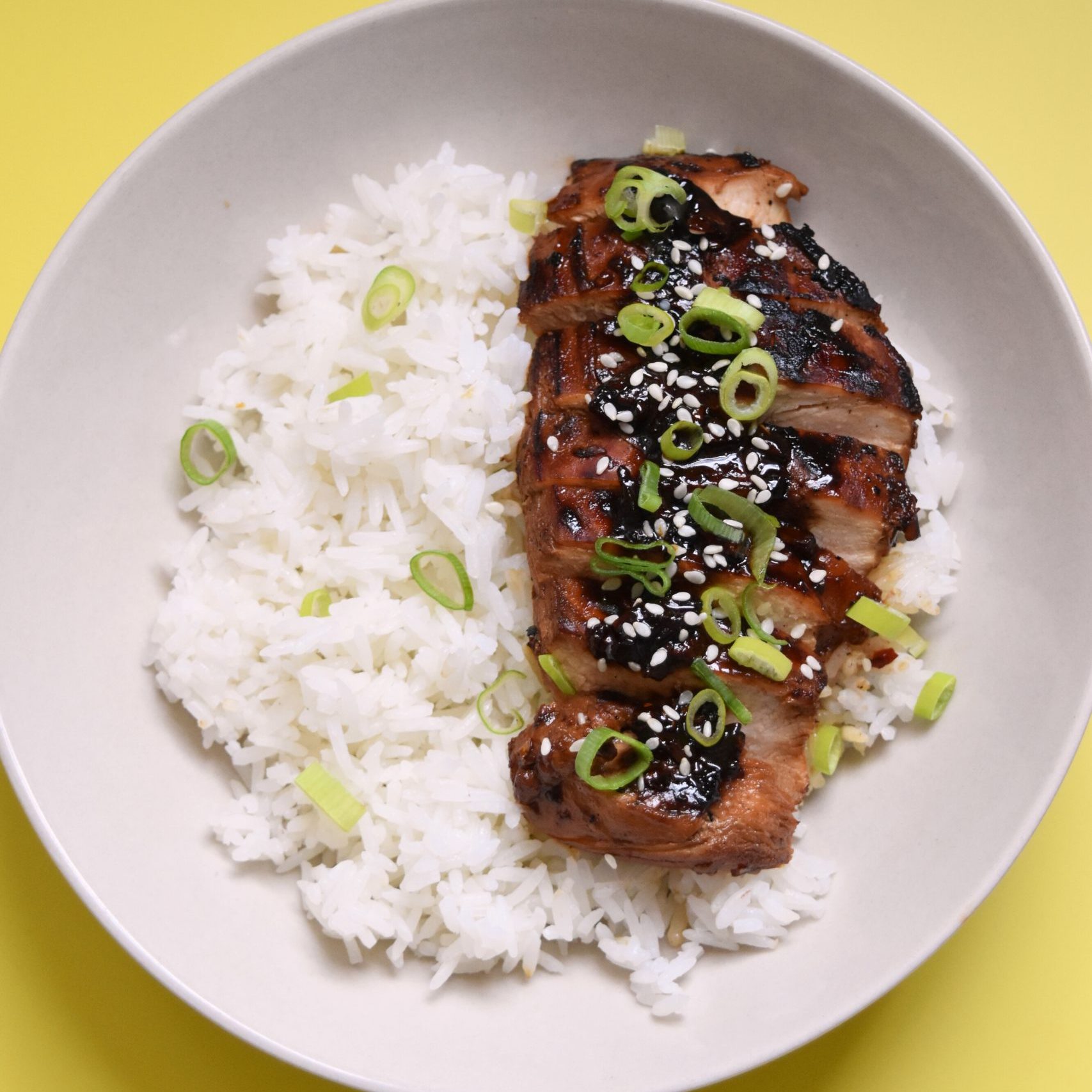 On a yellow backdrop, A plate of rice and honey soy chicken breast sliced up and garnished with sesame seeds and green onions.