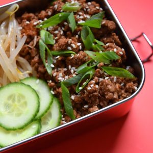 close up of korean beef with cucumber and beansprouts in a silver lunchbox on red background.