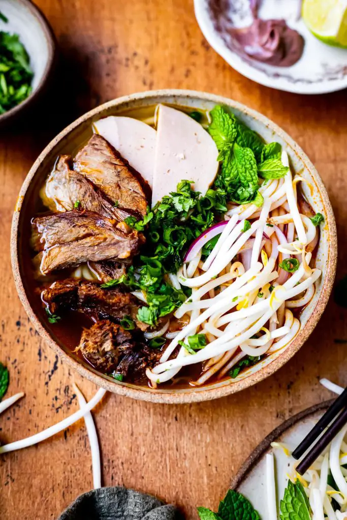 Best Vietnamese Soups: Birds eye view of bun bo hue. It is assembled and laid out perfectly, beansprouts, meat loaf (cha), brisket, greens for garnish on a wooden brown table. 