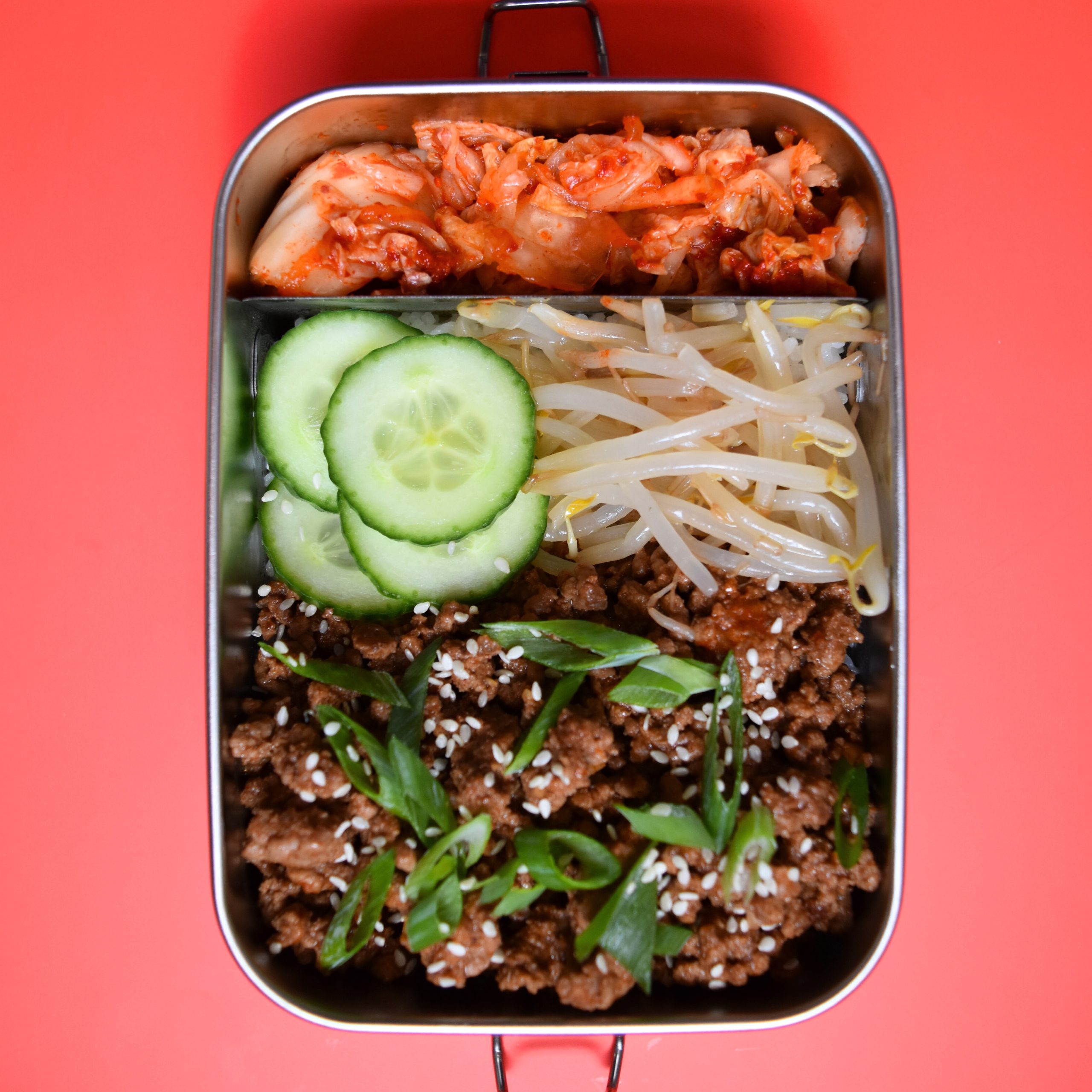 on red background, silver lunchbox with korean beef, cucumber, beansprouts and kimchi