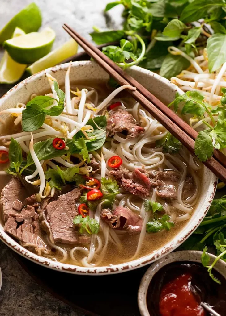 Best Vietnamese Soups: delicious pho in a bowl with wooden chopsticks resting on the bowl, garnishes such as lime, fresh herbs are in the background to show how pho should be eaten. 